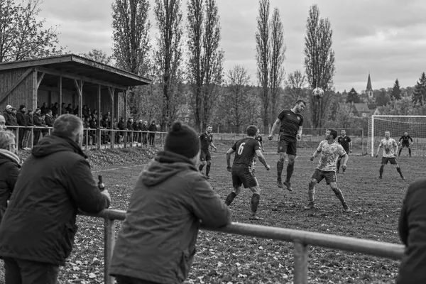 Kopfballduell auf einem matschigen Dorfsportplatz, Zuschauer am Geländer